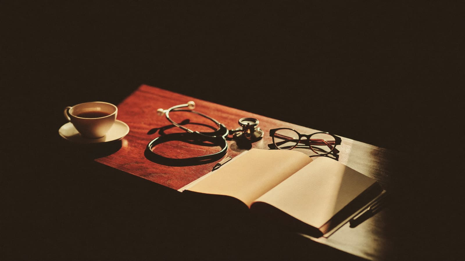 A stethoscope, reading glasses, and an open notebook in a shaft of warm light on a dark desk
