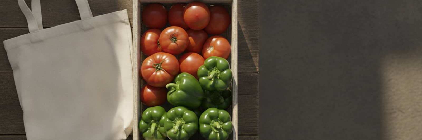 A canvas tote beside a wooden crate of ripe tomatoes and green bell peppers on a weathered outdoor table.