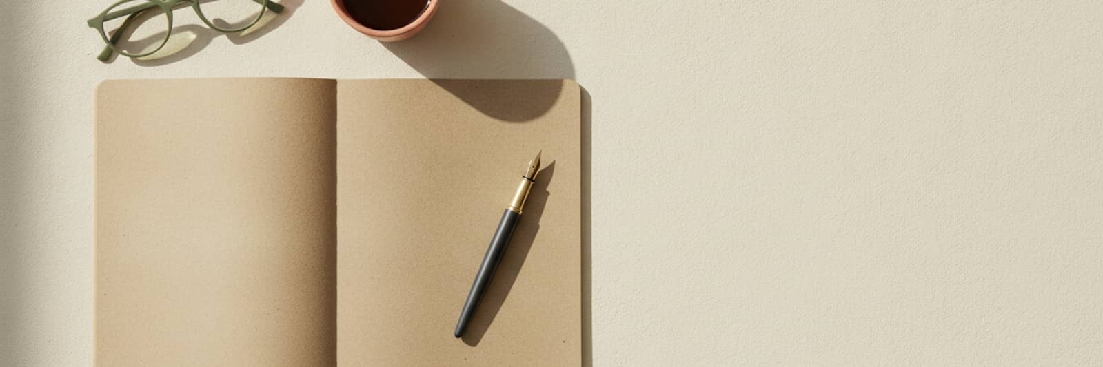 An open notebook with blank pages, a fountain pen, reading glasses, and a small cup of coffee on a pale desk.