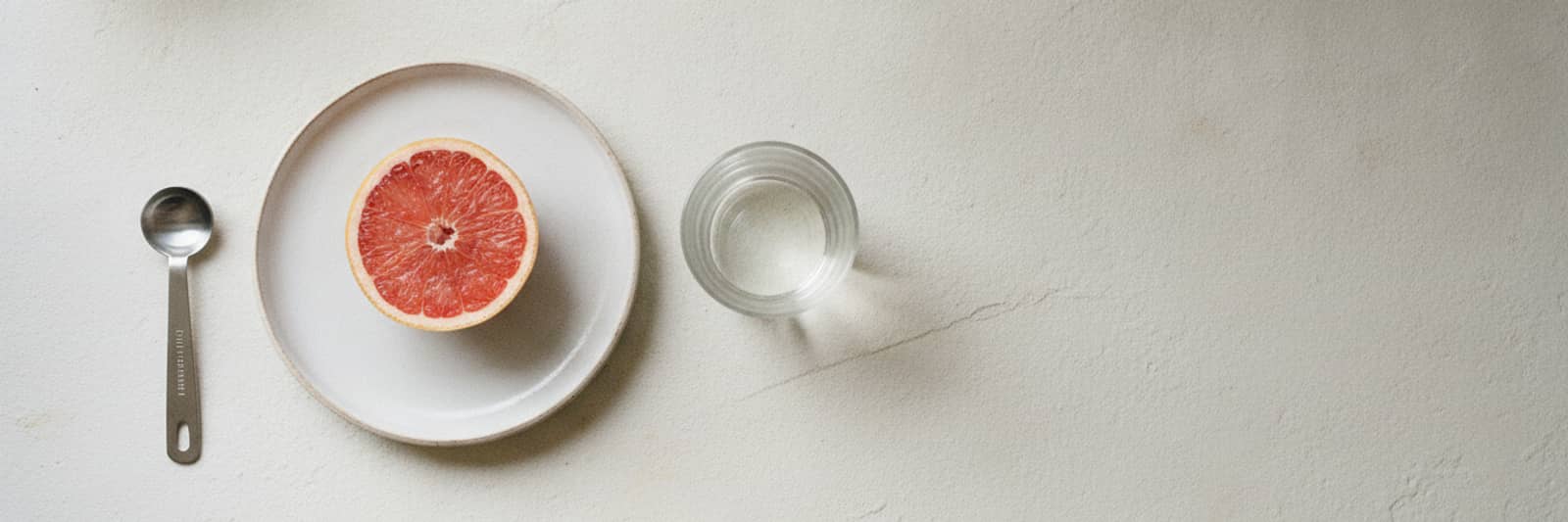A half grapefruit on a small plate, a glass of water, and a measuring spoon on a pale stone counter, seen from above.